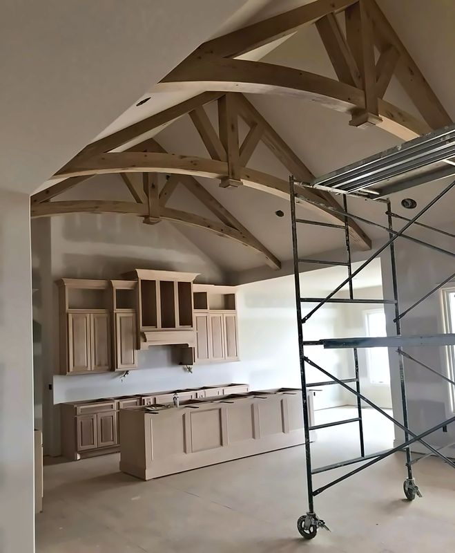 Arched rustic white oak timber trusses spanning a vaulted ceiling over a custom kitchen with cabinetry and a large island, before final finishes