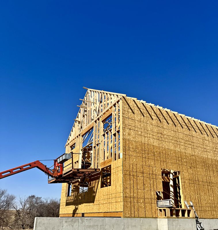 Two-story custom home framed and sheathed in OSB under a clear blue sky, with a telehandler lifting materials